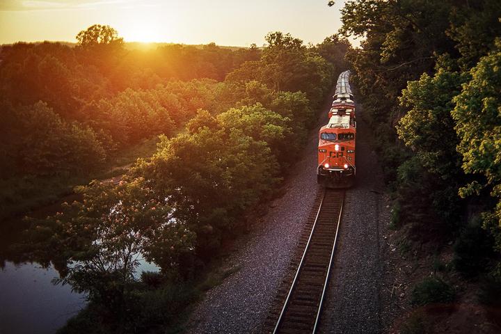 An orange train travels on a track through a forest at sunset.