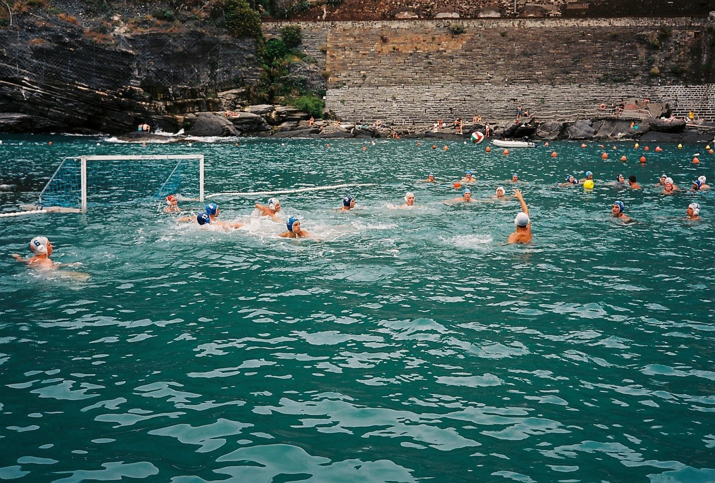 A group of people play water polo in the sea near a rocky shore.