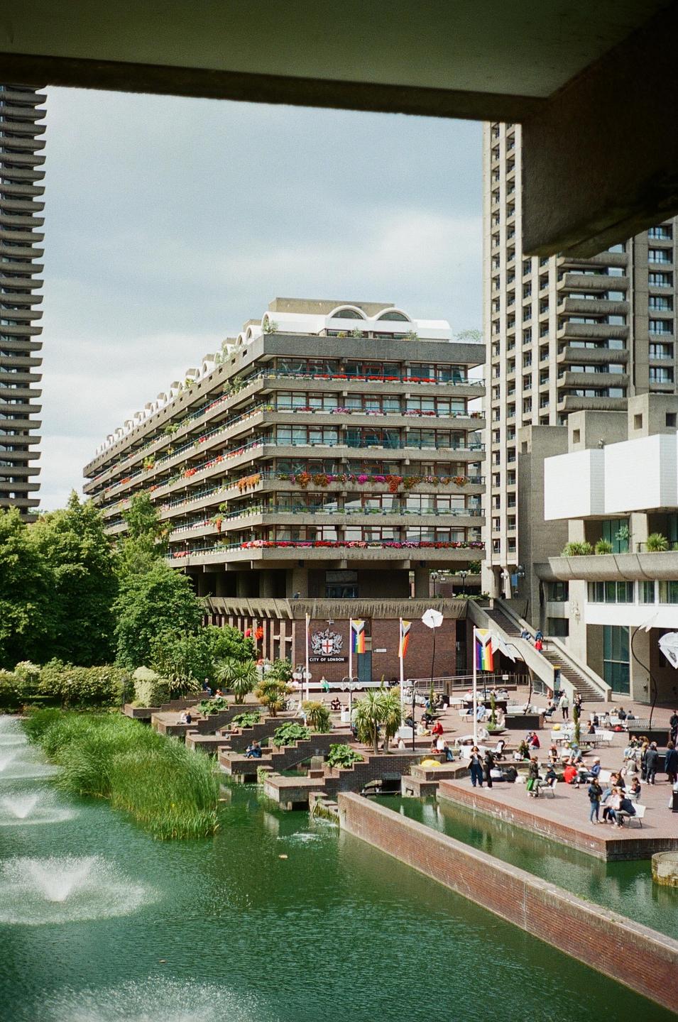 A brutalist building with balconies adorned with flowers overlooks a pond with fountains and people relaxing on a sunny day.