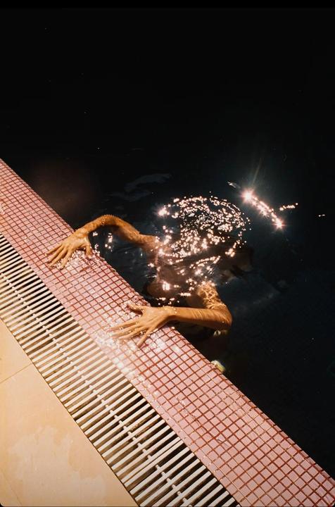 A person's arms and hands are visible in a dark swimming pool, holding onto the tiled edge with sparkling light reflecting on the water.