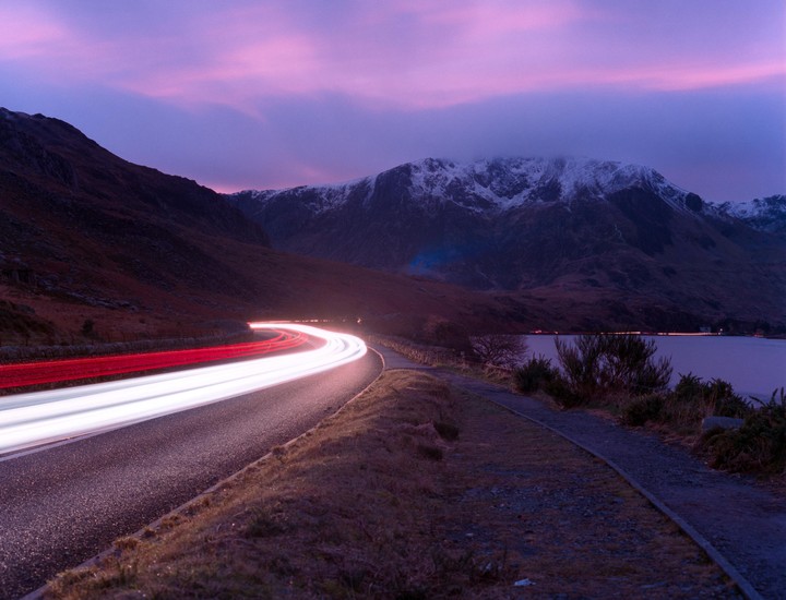 Light trails from vehicles illuminate a winding road next to a lake and snow-capped mountains under a purple sky.