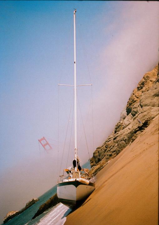 A sailboat is beached on a sandy shore next to a rocky cliff, with the Golden Gate Bridge visible through fog in the distance.