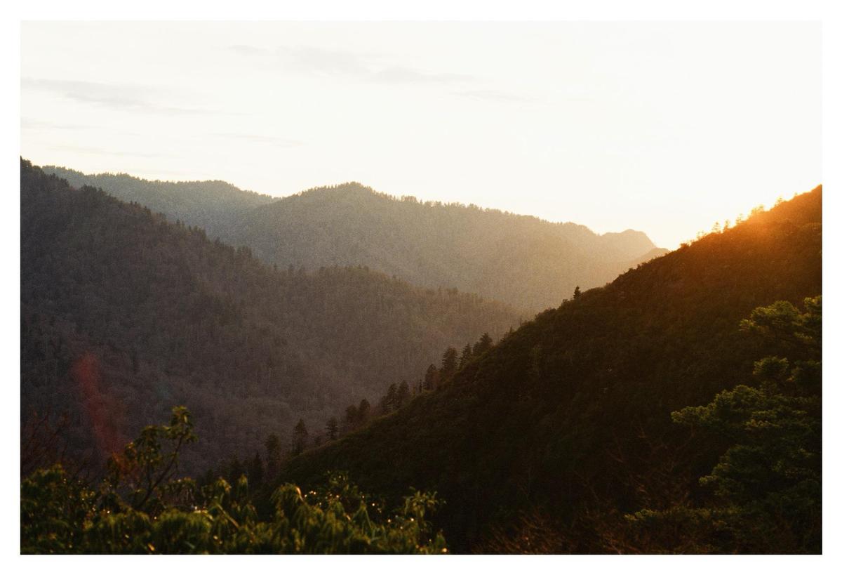 The sun sets over a mountain range covered in trees, with a bright lens flare on the right.