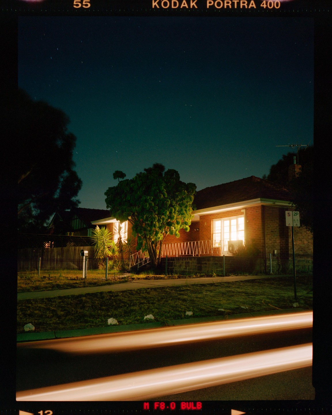 A brick house with illuminated windows sits under a starry night sky, with light trails from a vehicle passing in the foreground.