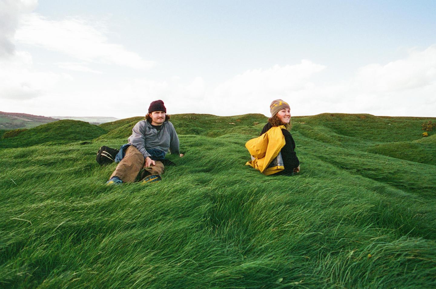 A man and a woman sit in a field of tall green grass under a cloudy sky.