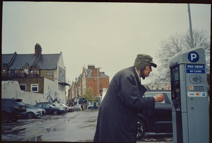 A man in a coat and hat stands next to a parking meter on a street with parked cars and buildings.