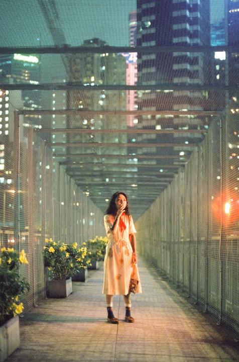 A woman stands on a pedestrian bridge at night, surrounded by city lights and potted plants.