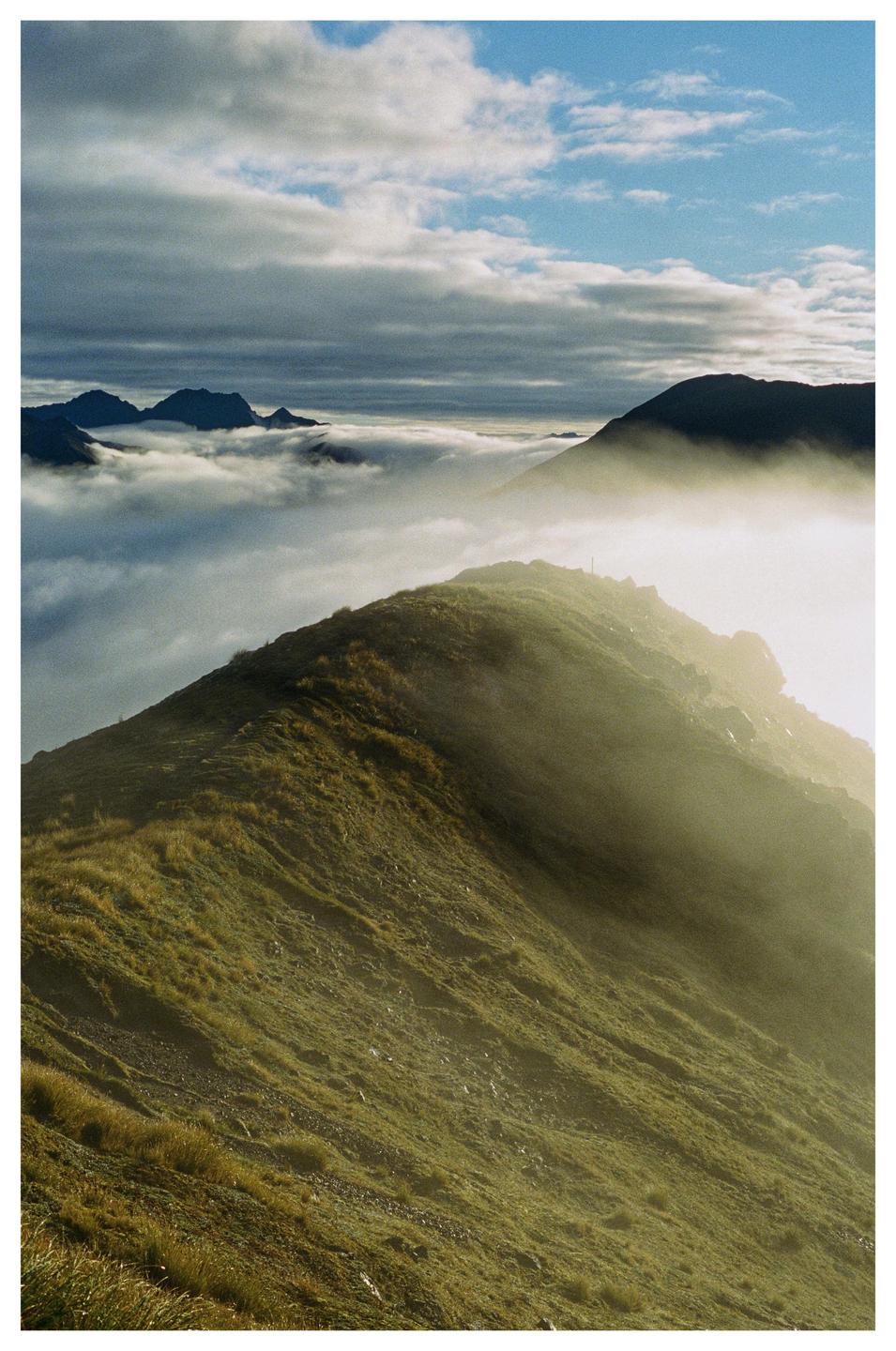 A mountain ridge covered in green grass is partially obscured by fog, with other mountain peaks and clouds visible in the distance.