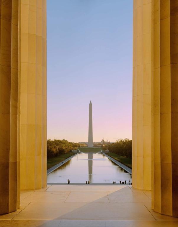 The Washington Monument is visible in the distance, reflected in the Lincoln Memorial Reflecting Pool, framed by two large columns.