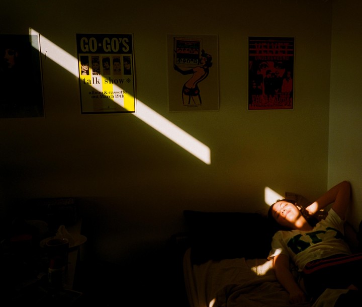 A woman lies on a bed in a sunlit room with posters on the wall.