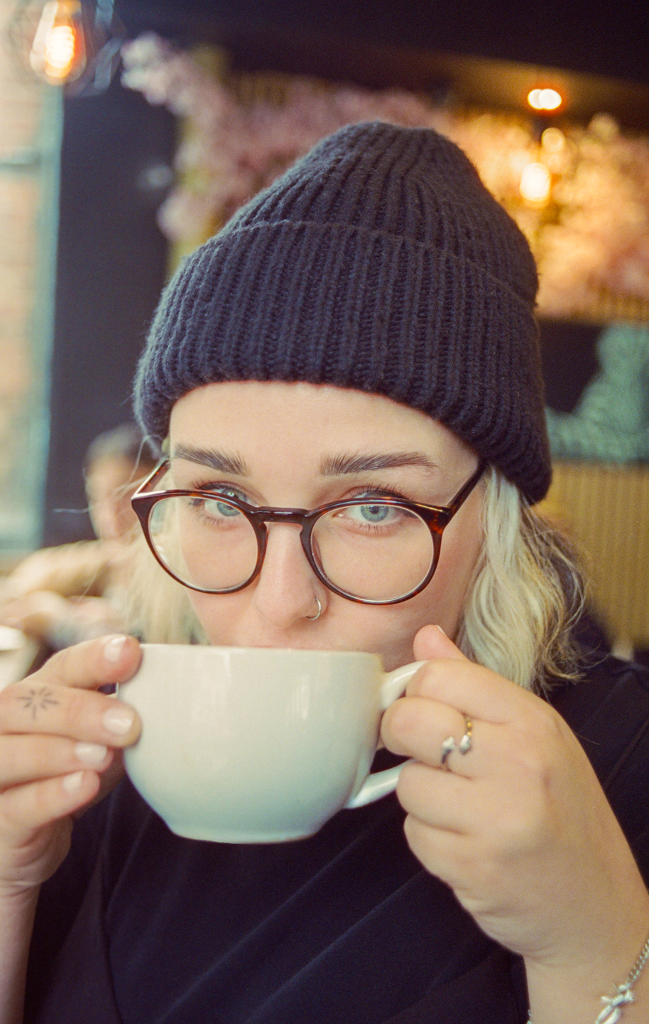 A woman wearing a beanie and glasses sips from a white mug.