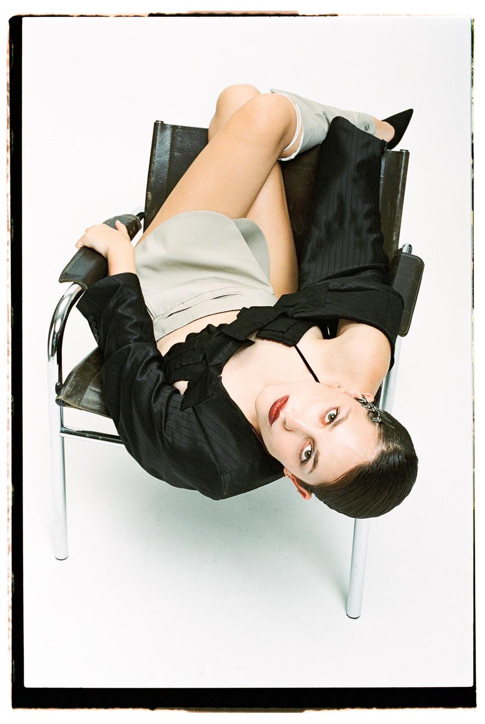 A woman with dark hair and red lipstick is lying upside down in a black leather chair, looking directly at the viewer.