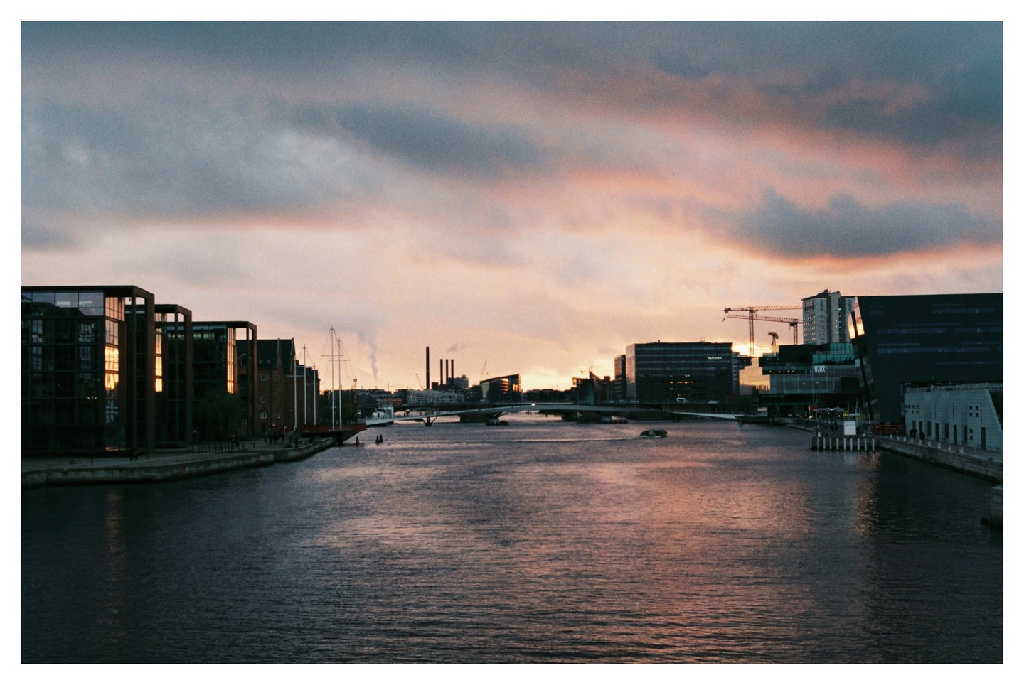 A city canal is lined with modern buildings and a sailboat under a dramatic sunset sky.