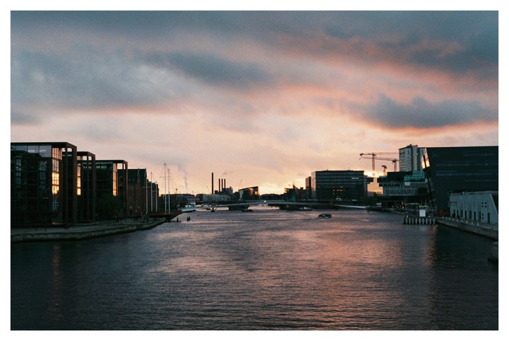 A city canal is lined with modern buildings and a sailboat under a dramatic sunset sky.