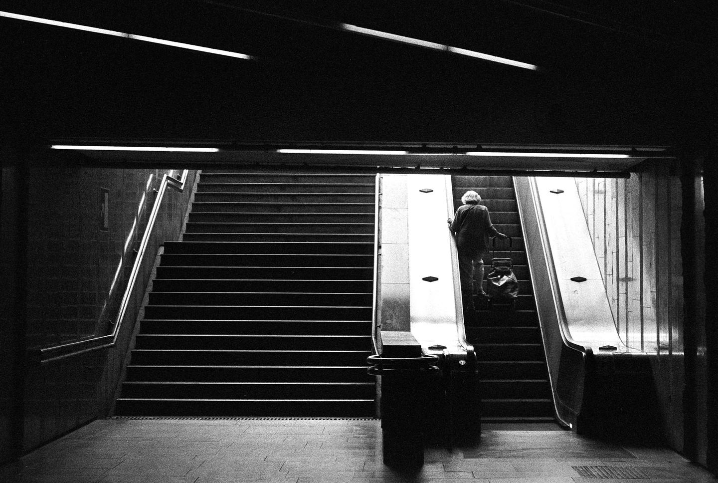 A woman with a shopping cart ascends an escalator next to a long staircase in a subway station.