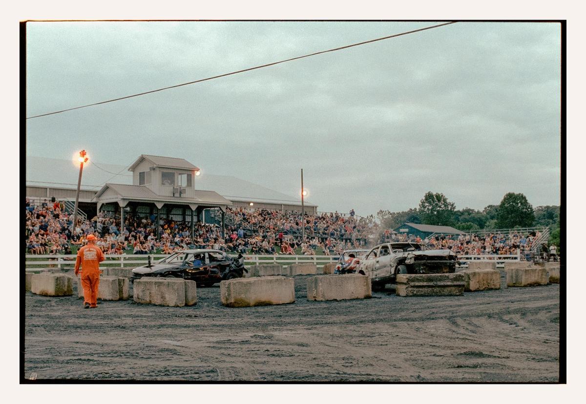 A man in an orange jumpsuit walks past wrecked cars in an arena with a crowd watching from bleachers.