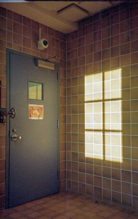 Sunlight streams through a window, casting a grid shadow on a tiled wall next to a closed door with a security camera above it.