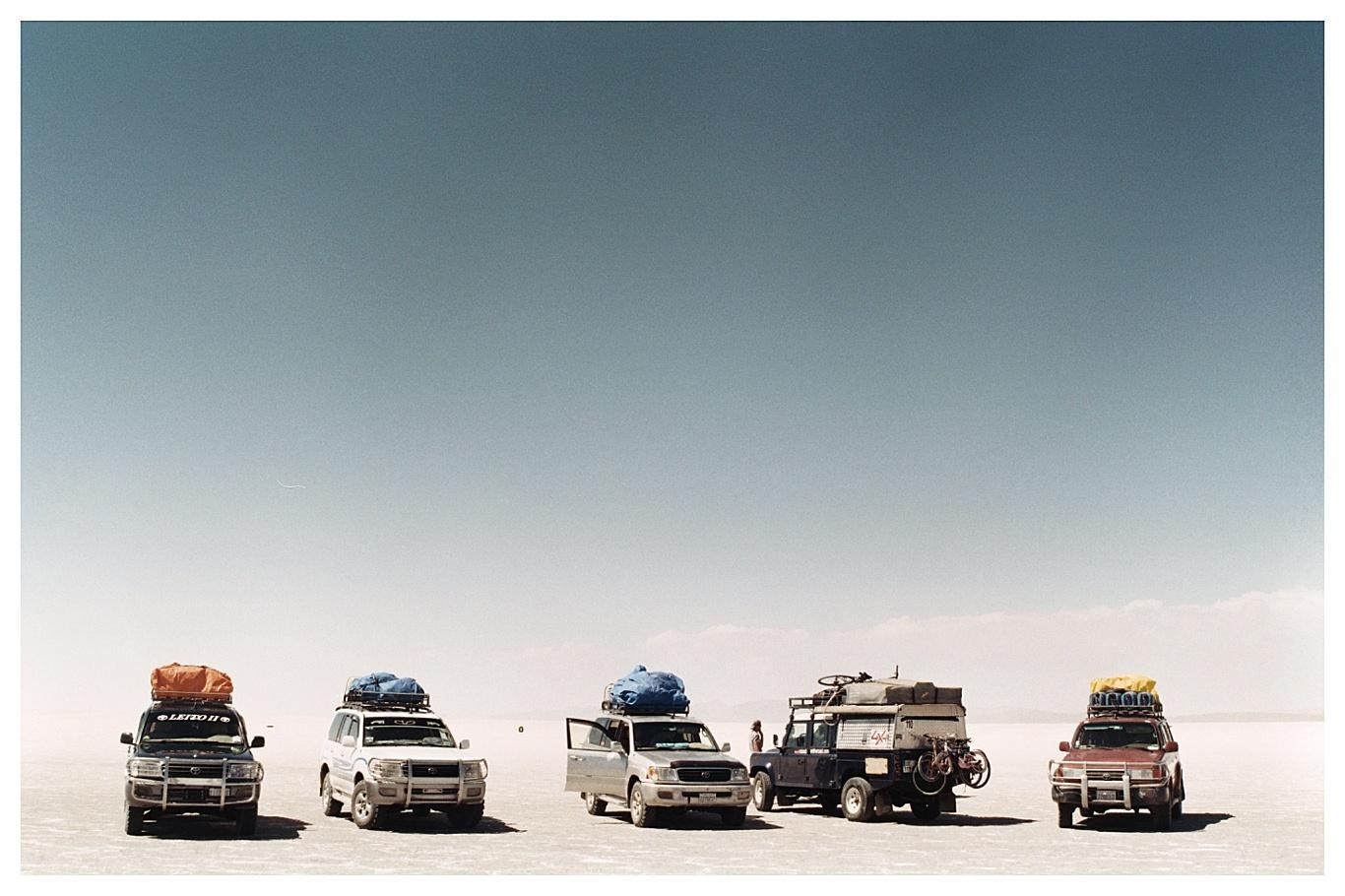 Five vehicles with gear on their roofs are parked on a vast, flat, and bright salt flat under a clear sky.
