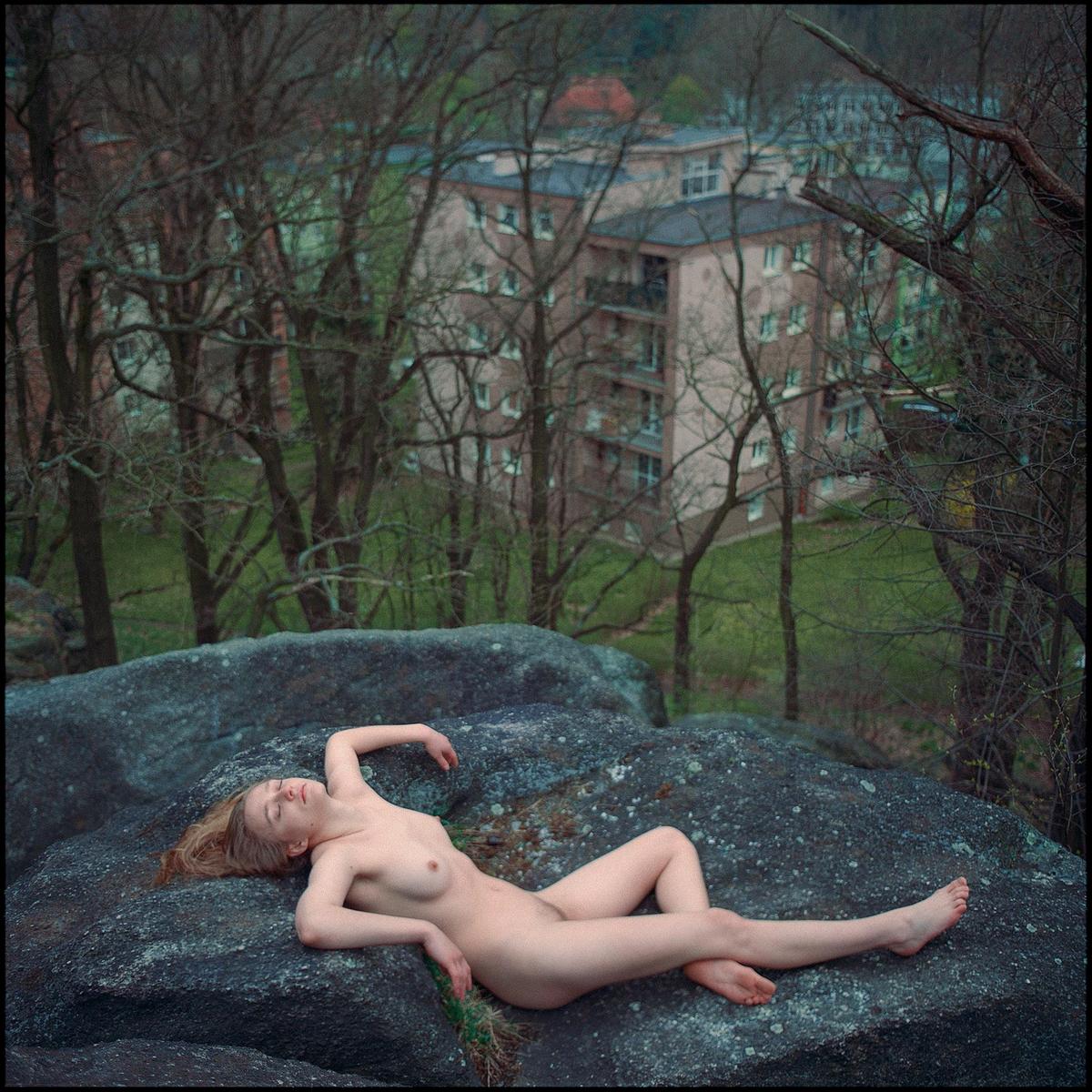 A nude woman lies on a large rock with buildings and trees in the background.