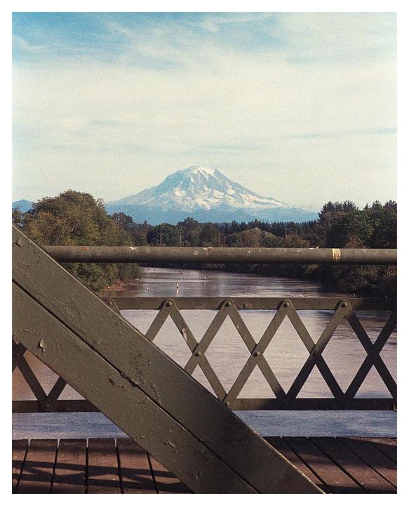 A snow-capped mountain rises in the distance behind a river and a bridge.