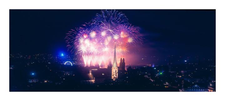 Fireworks explode over a city skyline at night, illuminating a tall church steeple and other buildings.