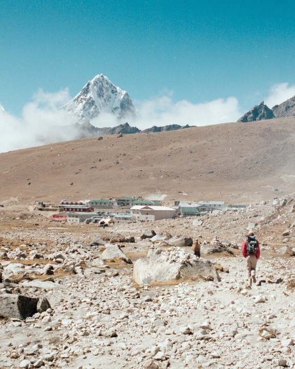 A person with a backpack walks towards a small settlement in a rocky, barren landscape with a snow-capped mountain in the distance.