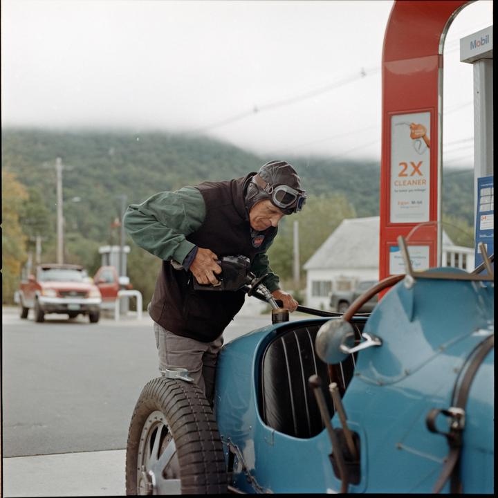 A man wearing a leather helmet and goggles fills a vintage blue car with gasoline at a gas station.