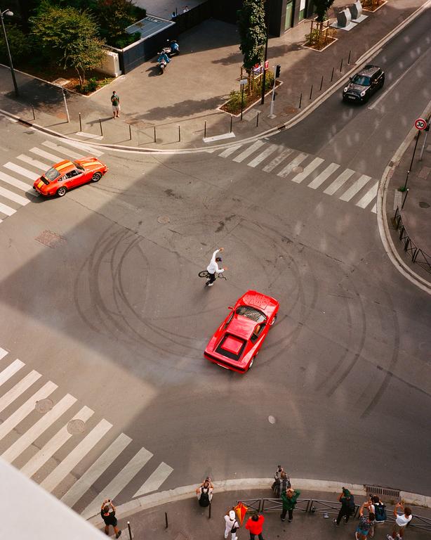 A red sports car and a cyclist are in the middle of a street intersection with tire marks on the asphalt, while people watch from the sidewalk.