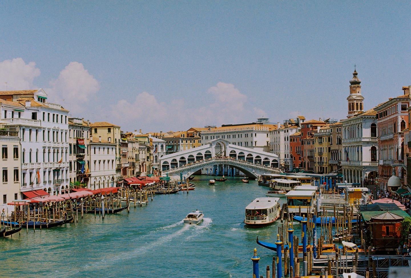 A wide shot shows the Rialto Bridge over the Grand Canal in Venice, with boats on the water and buildings lining the canal.