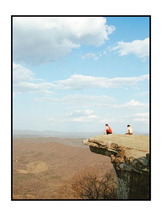 Two men sit on a rock outcrop overlooking a vast mountainous landscape under a cloudy sky.