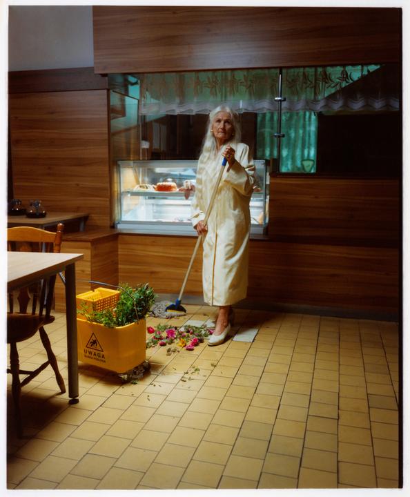 An older woman with long white hair sweeps a tiled floor in a room with wood paneling and a display case.