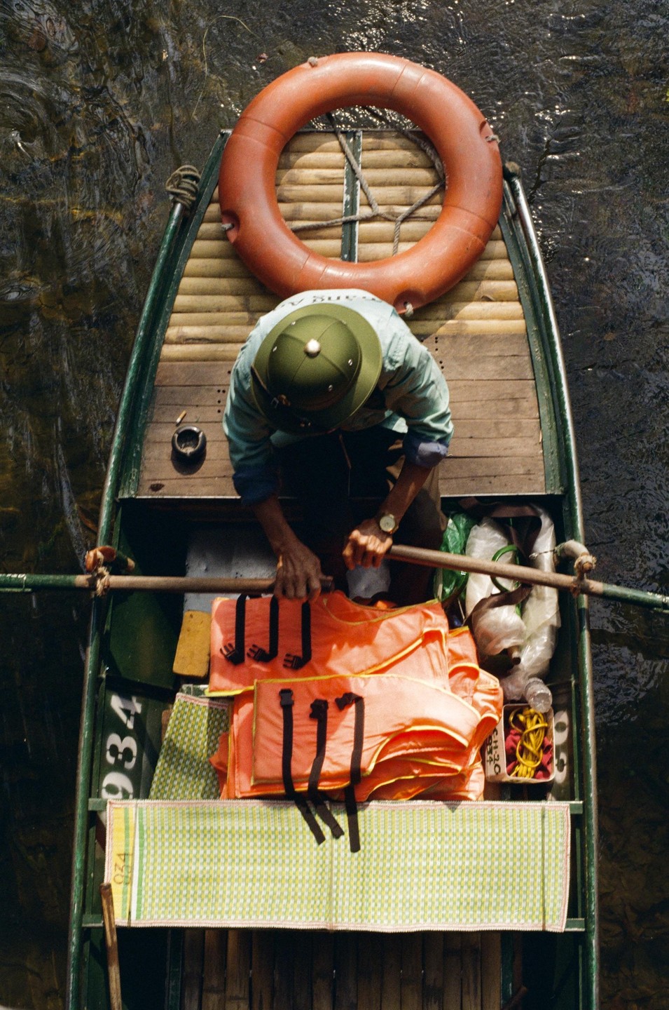 A person wearing a conical hat is seen from above, sitting in a wooden boat filled with orange life vests and a lifebuoy.