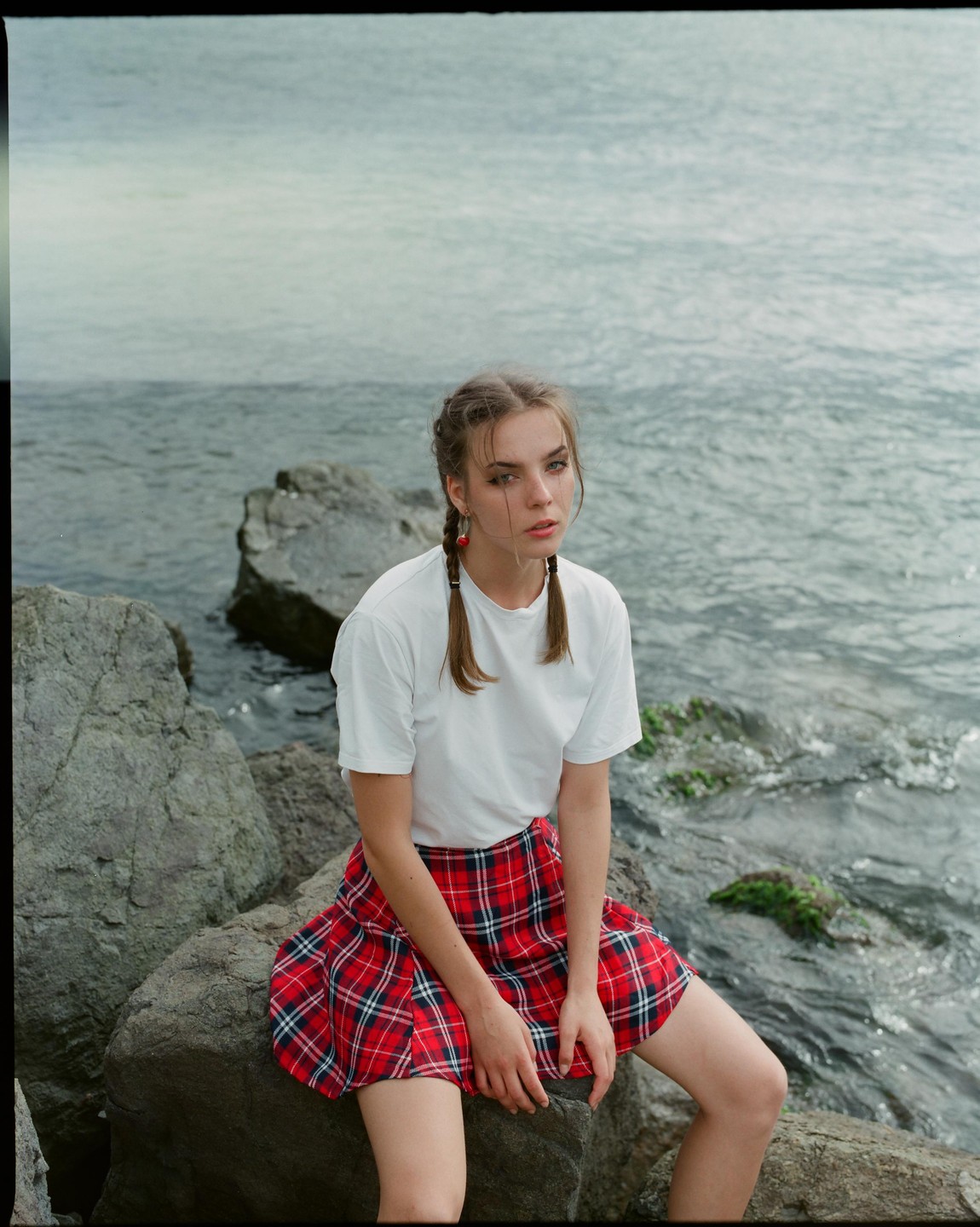 A young woman with braided hair and a red plaid skirt sits on rocks by the water.
