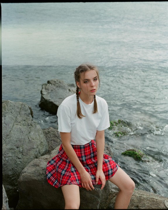 A young woman with braided hair and a red plaid skirt sits on rocks by the water.