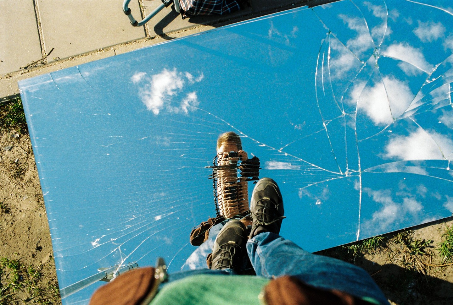 A man's reflection is visible in a broken mirror that reflects the sky and clouds.