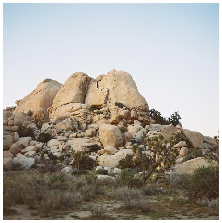 A large rock formation rises above desert brush and Joshua trees under a clear sky.