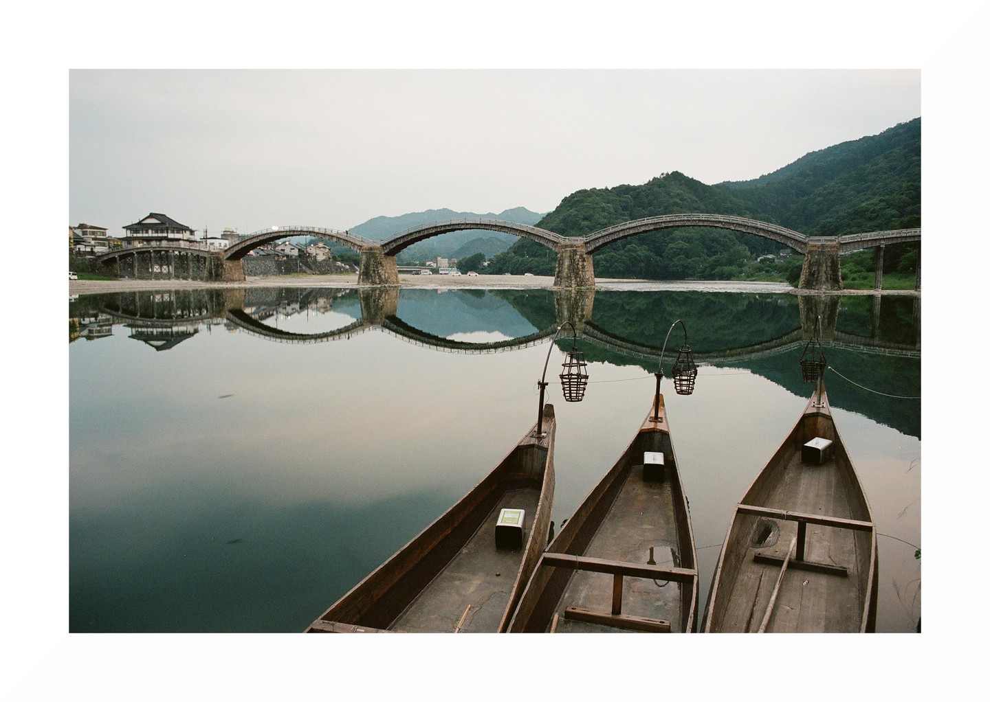 Three wooden boats float on a still river with a five-arched bridge and mountains in the background.