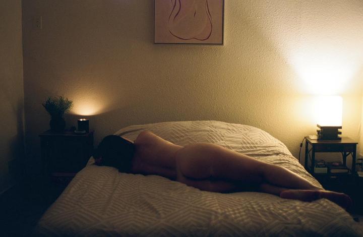 A woman lies nude on a bed in a dimly lit room.