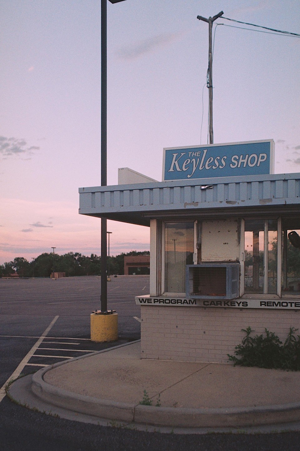 A small, abandoned shop with a sign that reads "The Keyless Shop" stands next to an empty parking lot under a pink sky.