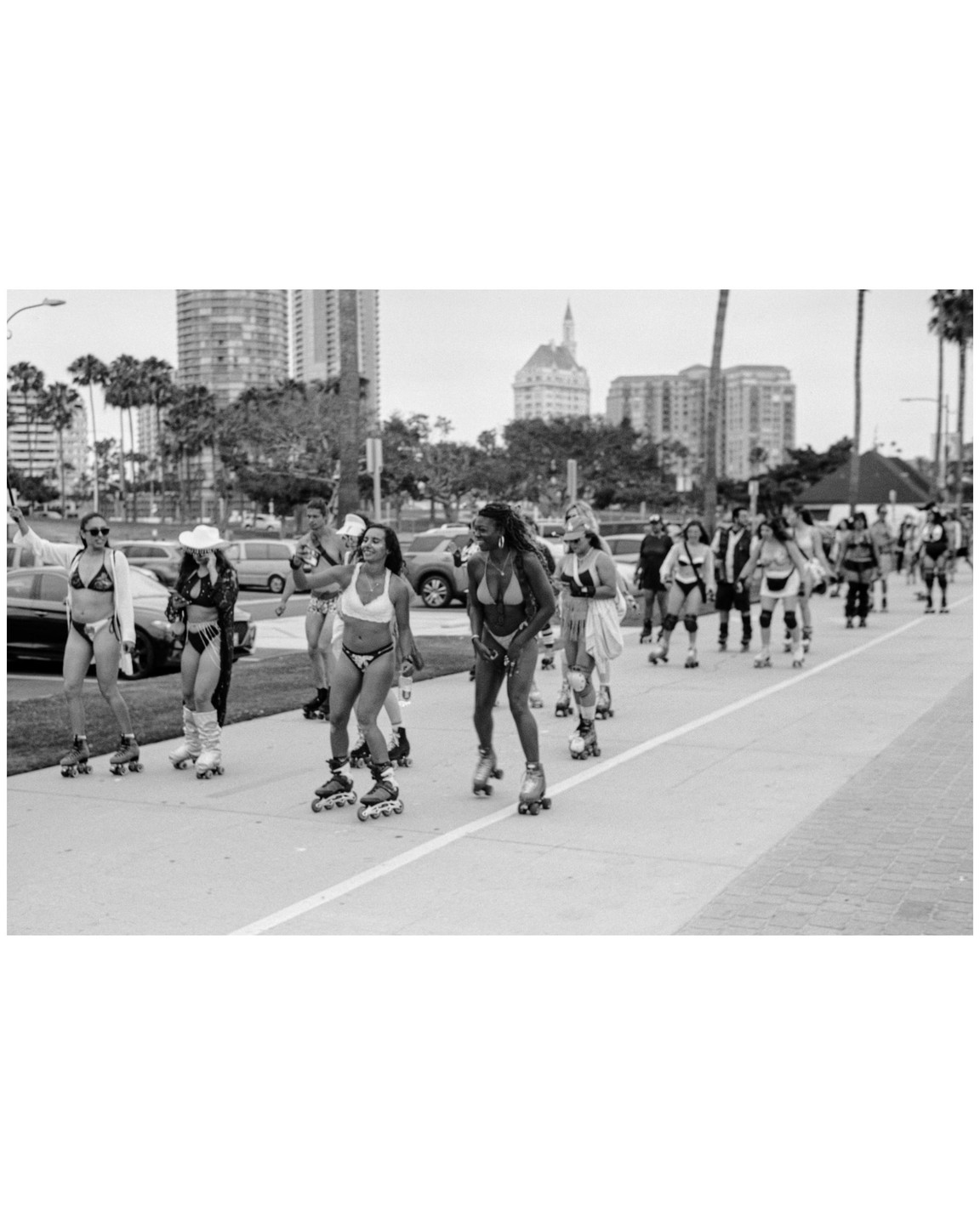 A group of women in bikinis and roller skates are moving along a paved path next to a street with buildings and palm trees in the background.
