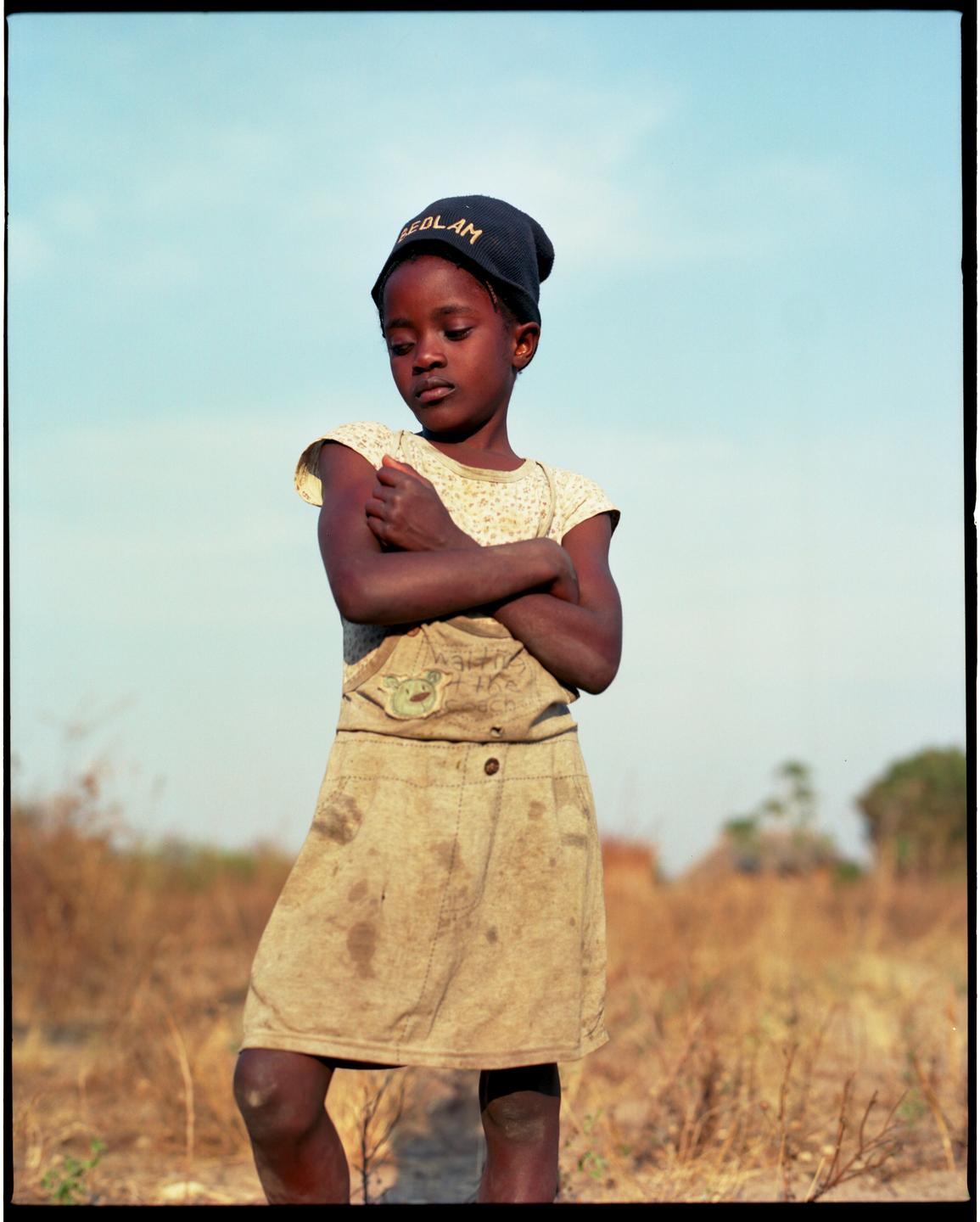 A young girl with a beanie and a dirty dress stands with her arms crossed in a field of dry grass.
