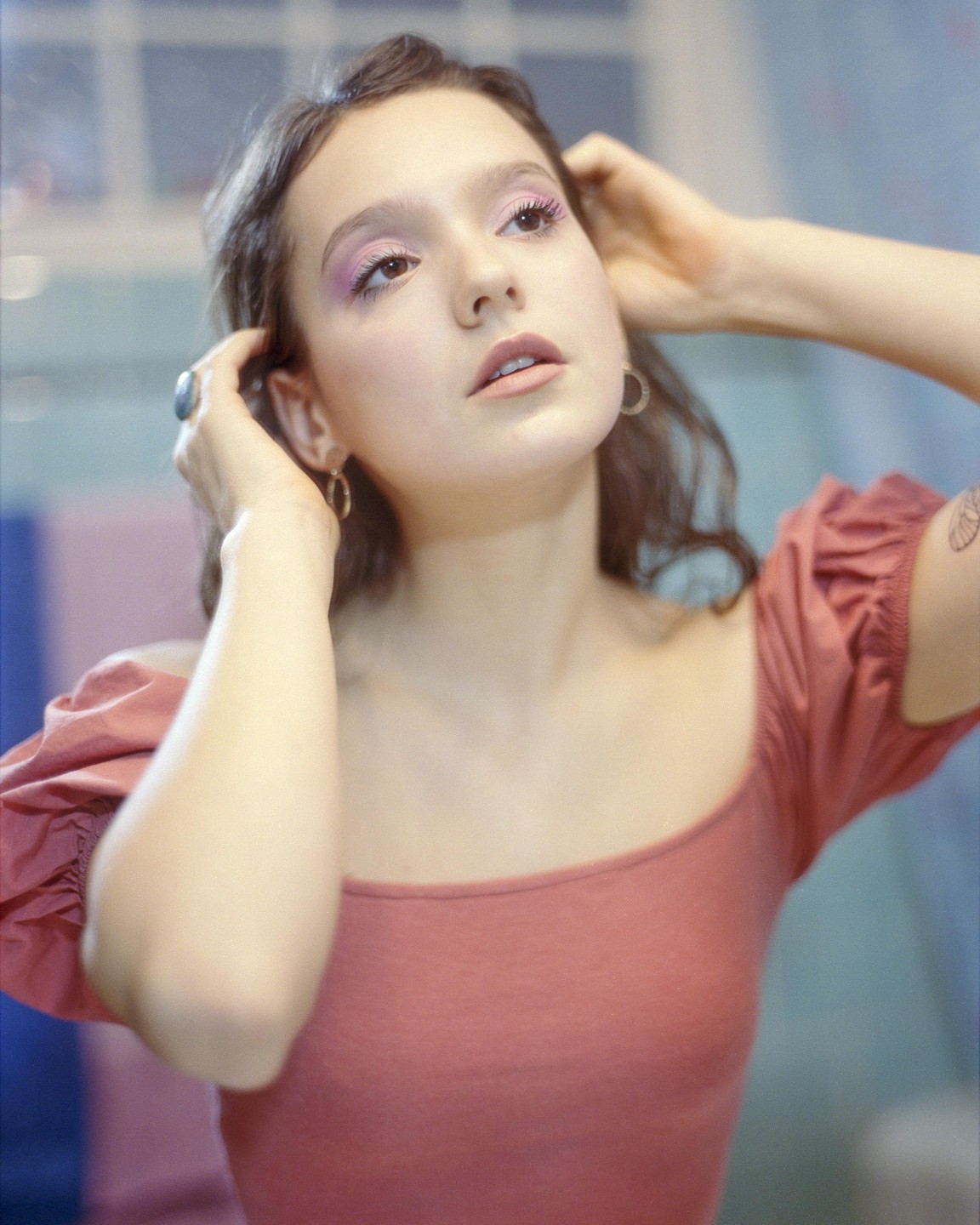 A woman with pink eyeshadow and a coral top looks up while holding her hair.