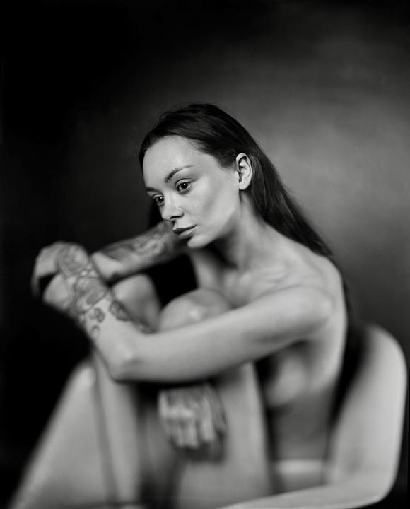 A woman with long hair and tattoos on her arms sits with her knees drawn up to her chest.