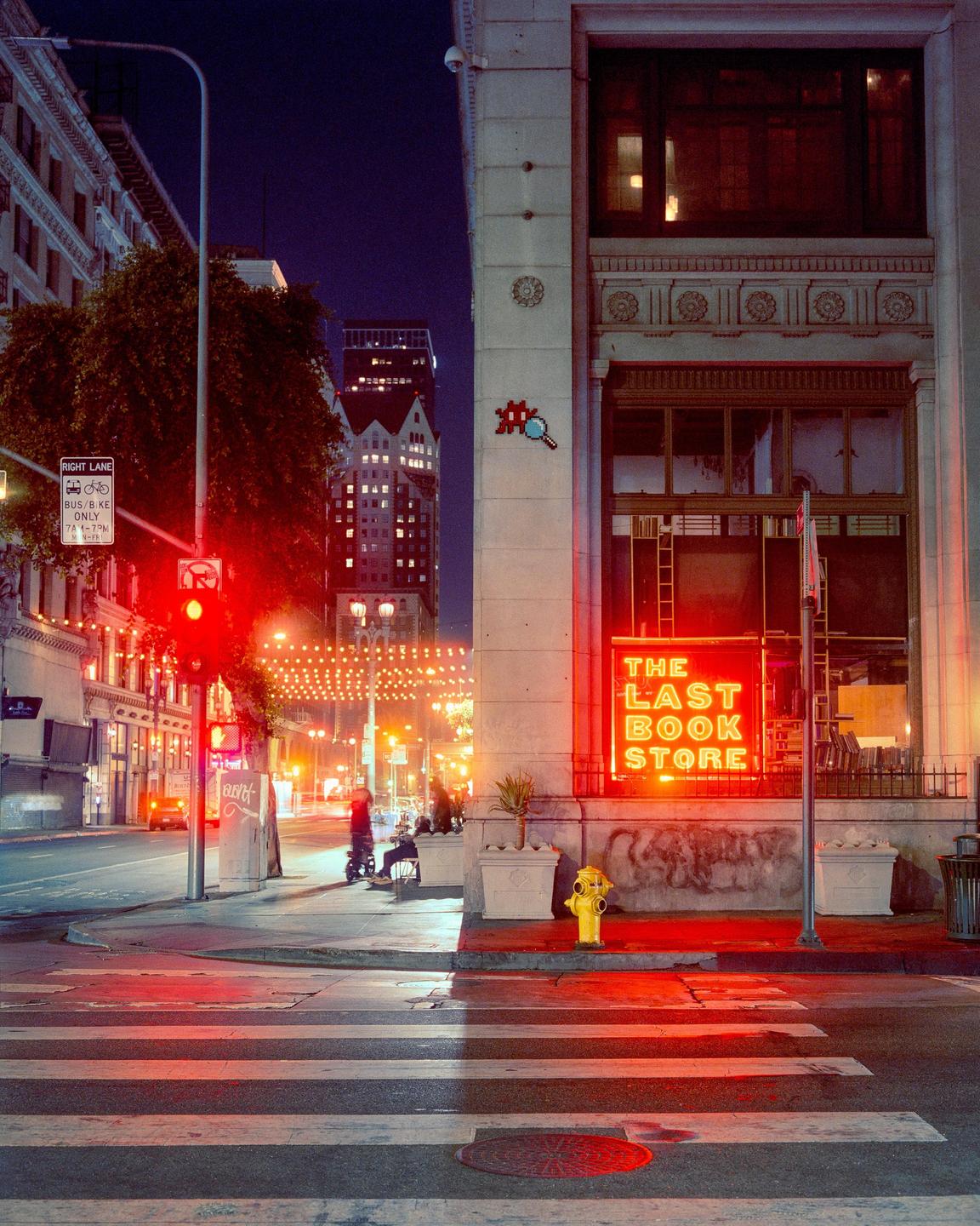 A street corner at night features a bookstore with a red neon sign, a fire hydrant, and a crosswalk.