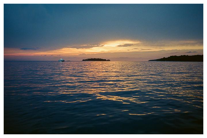 A boat with lights on is visible on the water with islands in the distance under a dramatic sunset sky.