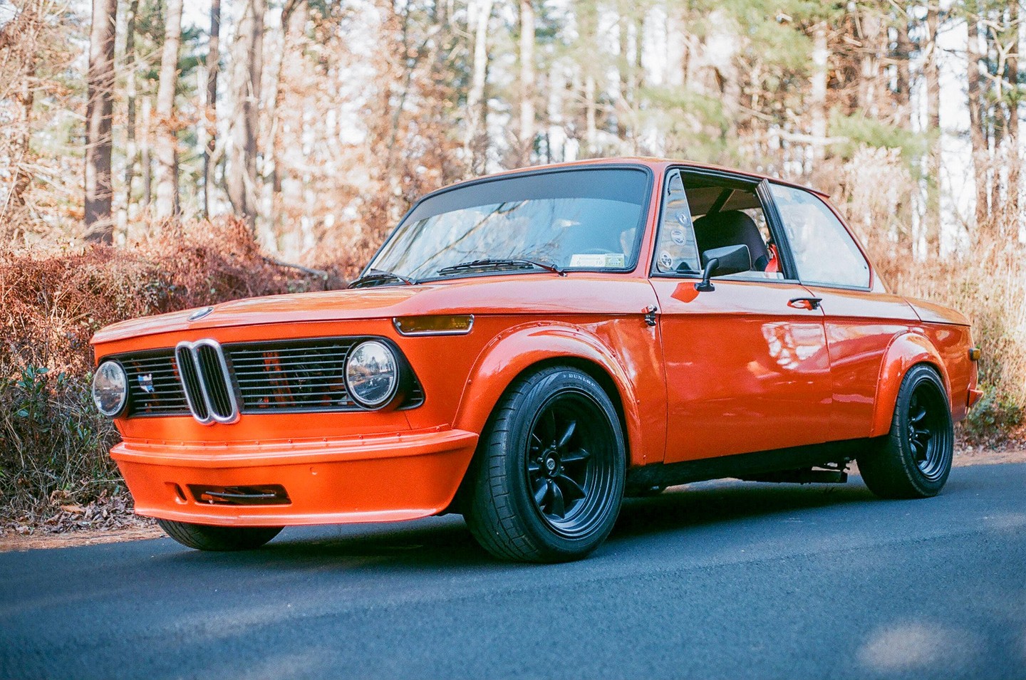 An orange vintage car with black wheels is parked on an asphalt road with trees and dry brush in the background.
