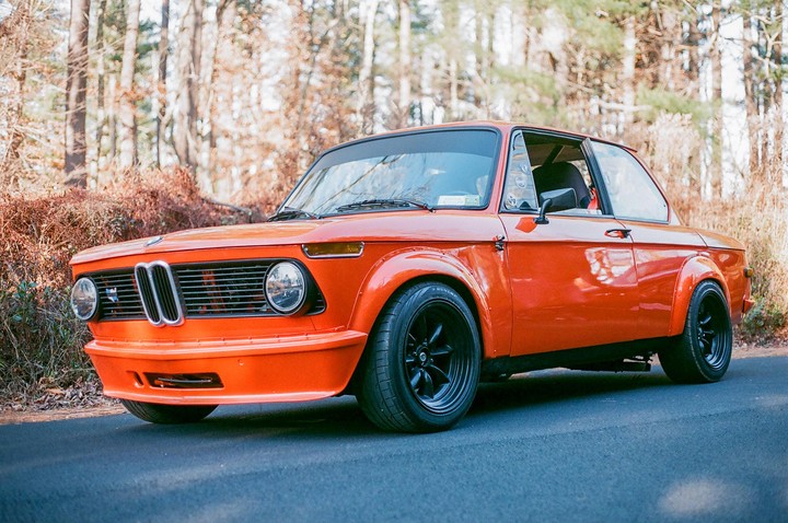 An orange vintage car with black wheels is parked on an asphalt road with trees and dry brush in the background.