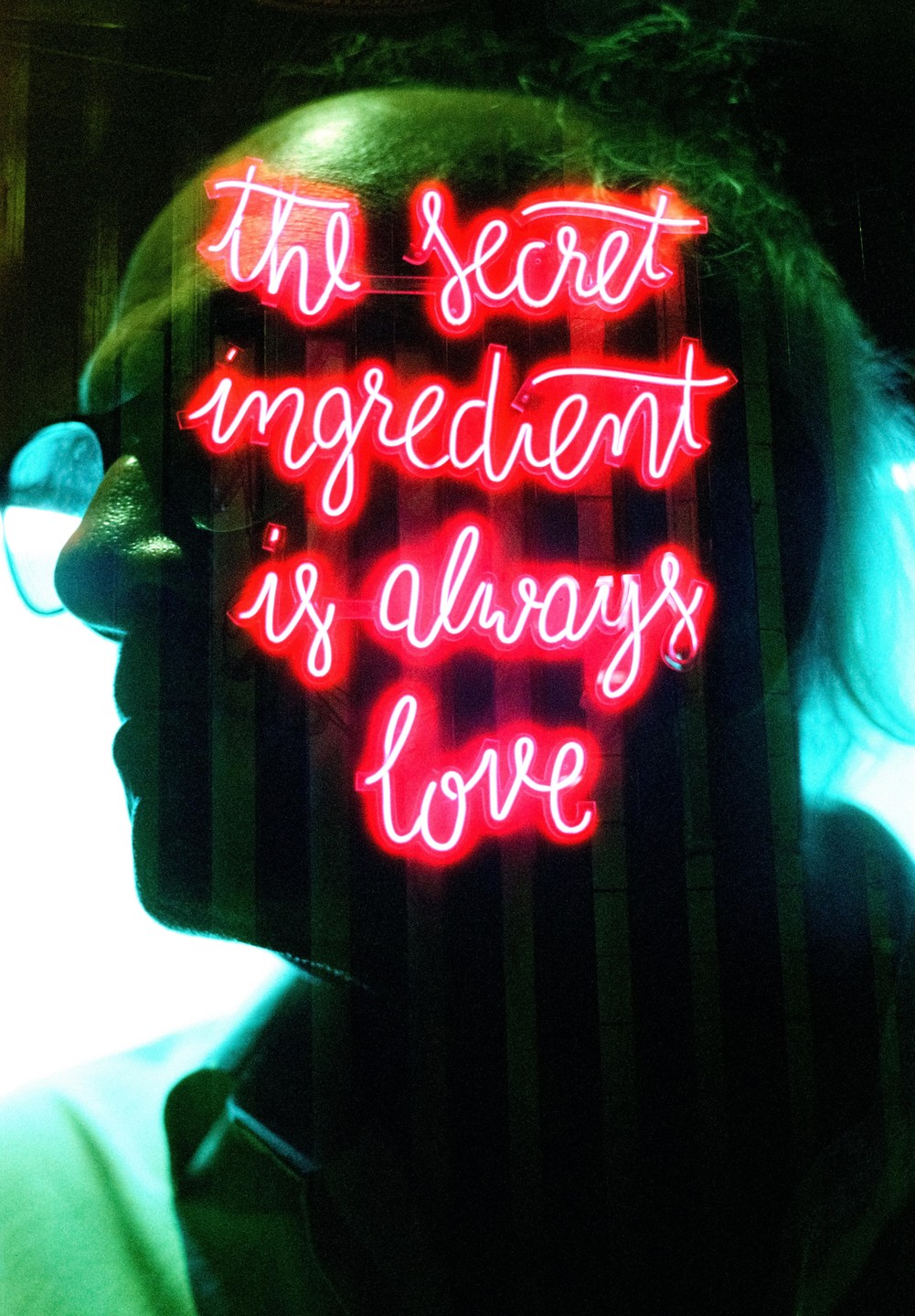 A neon sign with the words "the secret ingredient is always love" is double exposed over a man's profile.