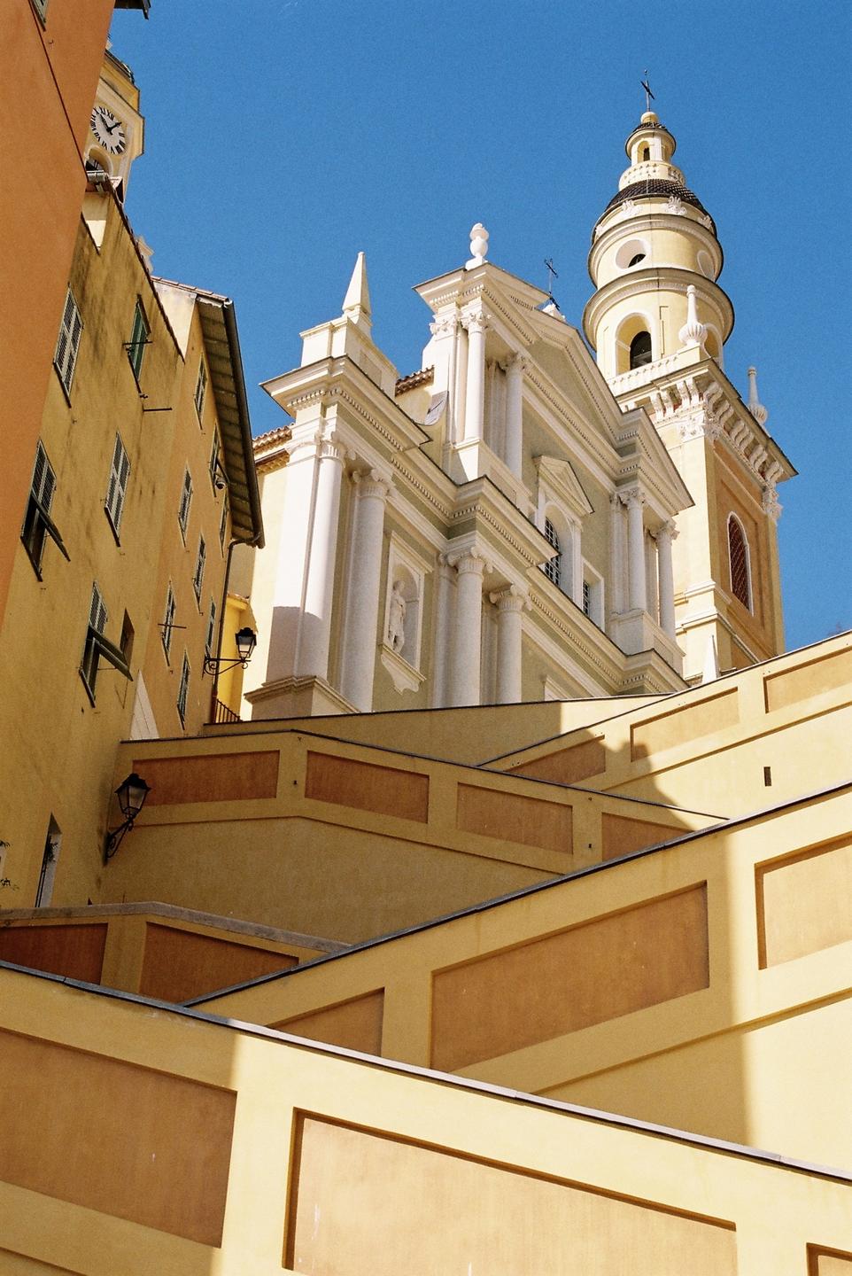 A low-angle view shows a yellow basilica with a bell tower and surrounding buildings under a clear blue sky.
