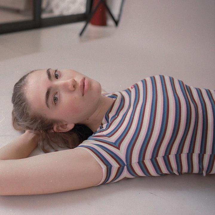 A woman with her hair pulled back lies on her back on a light surface, looking up at the viewer.